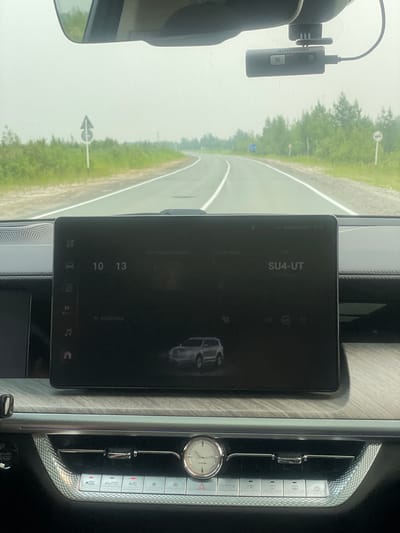 The image shows the interior of a vehicle on a road in Fedorovskii, Russia. The dashboard features a large touchscreen display, a digital clock showing 10:13, and a date of 25/06/2020. The car's environment controls are visible below the screen, including buttons for "AUTO," "A/C," and "MODE." A dashboard camera is mounted on the windshield. Outside the vehicle, a two-lane road stretches into the distance, bordered by green vegetation and trees. Road signs are visible on the side of the road. The sky is overcast, and the overall atmosphere suggests a hazy or misty condition.
