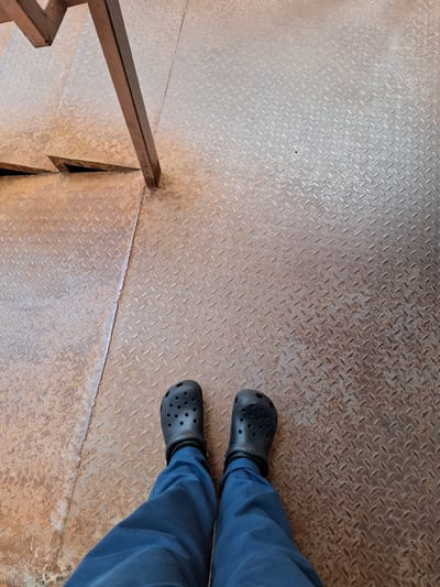 The image shows a pair of legs wearing blue pants and black Crocs, standing on a diamond plate metal surface. The surface appears to be part of a stairway, with a metal railing visible on the left side. The metal has a worn and slightly rusty appearance. The perspective is from above, looking down at the person's feet and the stairs. The lighting suggests it could be daytime, but there are no clear indicators of the weather or a specific time of day. The scene is simple and focuses on the footwear and the textured metal surface. There is no visible text in the image. The location context of Grigorovo, Russia, is provided but not visually evident from the image itself.
