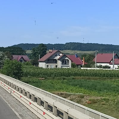 The image depicts a rural scene with several houses in the background, situated amidst greenery and a cornfield. In the foreground, there is a bridge or overpass with a white railing. The sky is clear and blue. The houses appear to be modern, with red tiled roofs. One house is particularly prominent, featuring a complex roofline with multiple gables and triangular windows. To the right, another house with a red roof and white walls is visible. The surrounding landscape is lush, with trees and crops. In the distance, hills can be seen. The overall impression is one of a tranquil countryside setting.