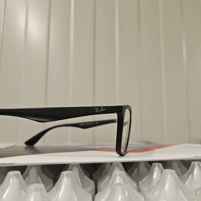 The image shows a close-up of a pair of black Ray-Ban eyeglasses resting on a white surface. The background consists of vertical, light-colored panels, likely a wall. The eyeglasses are positioned horizontally across the frame, with the left temple in focus and the right lens slightly out of focus. The Ray-Ban logo is visible on the left temple. Below the eyeglasses, a sliver of what appears to be paper or packaging with a red stripe is visible. The bottom of the image reveals a row of white, translucent plastic caps, arranged in a grid pattern. The scene is static and does not depict any action or interaction. The location in Nowa Wieś Książęca, Poland, is not discernible from the visual information provided in the image.