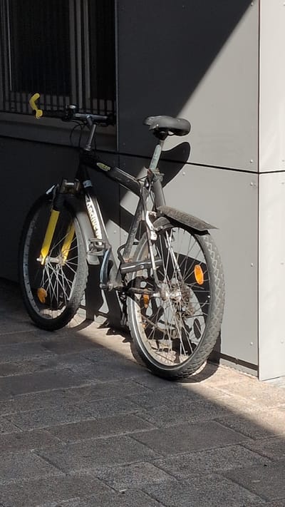 A black bicycle with yellow accents is parked on a brick sidewalk next to a grey building. The bicycle has a black frame, yellow front fork, black handlebars with a yellow grip, and a black seat. The front wheel is partially visible, and the rear wheel is prominently displayed. The bicycle appears to be a mountain bike. The building has a modern, grey facade with vertical panels and a window with vertical bars. Sunlight casts a shadow from the building onto the sidewalk and the bicycle. The scene is outdoors, and the lighting suggests it is daytime. The image does not provide enough information to determine the specific location within Osnabrück, Germany.