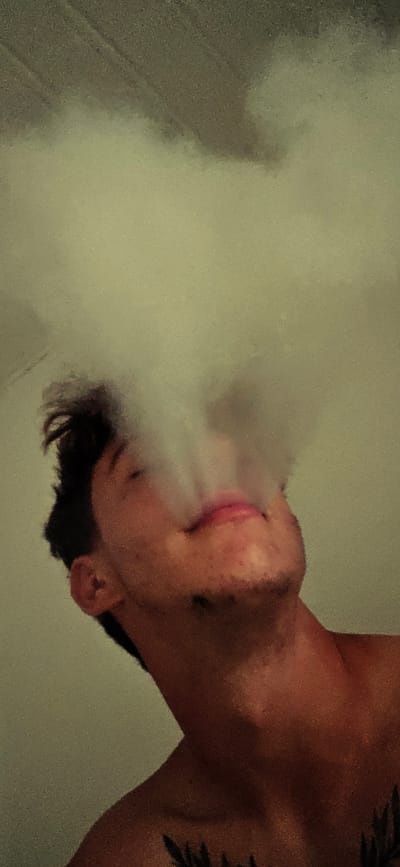 A low-angle, close-up, vertical shot shows a young person's face and upper chest, illuminated by soft, diffused light. They are looking upwards, with their eyes closed and a slight smile on their lips. A plume of white vapor, possibly smoke, is rising from their mouth and obscuring the upper part of their face and the background. Their hair is dark and appears somewhat tousled. The skin tone is fair, with subtle red undertones. A dark tattoo is visible on their upper chest. The background is a plain, light-colored wall, with some darker, textured areas at the top that might suggest a ceiling or a wall feature. The overall impression is intimate and serene. The image provides no information that would allow for its location in Kitzscher, Germany, or any other specific geographical location.