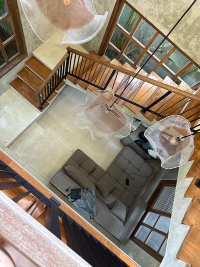 An overhead shot captures the interior of a multi-story building, likely a residential space in Payangan, Indonesia, featuring a contemporary design with concrete walls and wooden accents. A prominent wooden staircase with black metal railings ascends from the ground floor. On the ground level, a large, grey sectional sofa is positioned near the base of the stairs. Visible on the sofa are a remote control and a black pouch. To the left of the sofa, a grey jacket is draped over a surface. Shoes, including black boots, a pair of green shoes, and white sneakers, are placed near the sofa. Two unique, bell-shaped chandeliers with white mesh shades and visible bulbs hang from the ceiling, illuminating the space. One chandelier is positioned above the sofa, and another is suspended over the staircase. A large window with wooden frames overlooks an outdoor area with lush greenery and tree branches, suggesting a tropical environment. The flooring is a combination of polished concrete and light-colored tiles. The scene appears to be a moment of stillness, with no individuals actively engaged in any discernible actions, though a pair of bare feet can be seen on the lower steps of the staircase, suggesting someone is present but not fully visible.