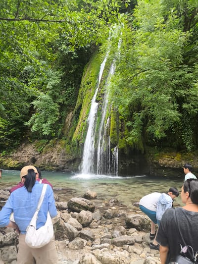 The image depicts a waterfall in Pirveli Balda, Georgia. A multi-tiered waterfall cascades down a moss-covered rock face into a clear, shallow pool. Several people are visible near the water's edge and in the foreground. In the foreground, a person in a blue shirt and tan pants carries a white crossbody bag. To their right, another person in a dark shirt faces away from the camera. To the left of the main foreground figure, another person in a burgundy top is partially visible. In the midground, a young person wearing a white shirt and blue shorts bends down, appearing to examine the rocks. Further back, on the right side of the image, a young person in a light green shirt stands looking towards the waterfall. The surrounding environment is lush with green foliage and trees, with large rocks scattered along the water's edge. The water is transparent, revealing the rocky riverbed.
