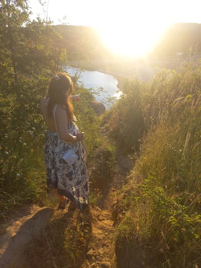 A woman in a long, patterned dress and sandals stands on a dirt path overlooking a body of water. She has long, dark hair and is holding a light blue camera bag. The sun is setting, casting a warm glow over the scene, with a bright sunburst in the upper right corner. Lush green vegetation, including tall grasses and trees, surrounds the path and the water. The location is likely near the coast, possibly at a viewpoint overlooking the Black Sea or a river in Sinemorets, Bulgaria, given the natural landscape and lighting conditions indicative of a sunset.