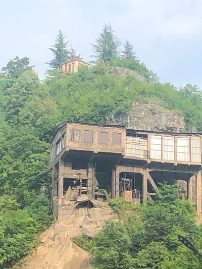The image shows an old, abandoned industrial building nestled on a hillside in Chiatura, Georgia. The building appears to be constructed of concrete and brick, with large windows that are partially boarded up. The structure is multi-leveled and supported by concrete pillars, suggesting it might have been a former mine or processing facility. The surrounding environment is lush and green, with dense foliage and trees covering the hillside. In the background, higher up on the hill, another smaller building with a red roof is visible, and a cableway system is suggested by thin lines extending across the sky. The overall impression is one of industrial decay set against a natural, verdant landscape. There are no people or active processes depicted in the image.