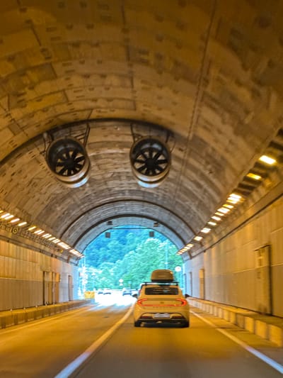 The image shows the interior of a tunnel. Two large circular fans are visible on the arched ceiling of the tunnel, resembling eyes. The tunnel walls are made of concrete with visible seams and reinforced concrete structures. Lights are installed along the sides of the tunnel ceiling, illuminating the roadway.
In the foreground, a yellow-white car with a roof box is driving through the tunnel. The car's rear is visible, and it has a license plate. Other cars can be seen in the distance, driving towards the exit of the tunnel. The exit of the tunnel reveals a bright outdoor scene with green trees and a blue sky. The roadway is asphalt, with white lane markings. The overall impression is of a modern tunnel infrastructure.
There are no people visible in the image. The context of Monastyr, Russia, is not discernible from the visual elements of the image itself. The scene depicts a transportation artery designed to facilitate movement through a mountainous or otherwise obstructed terrain.