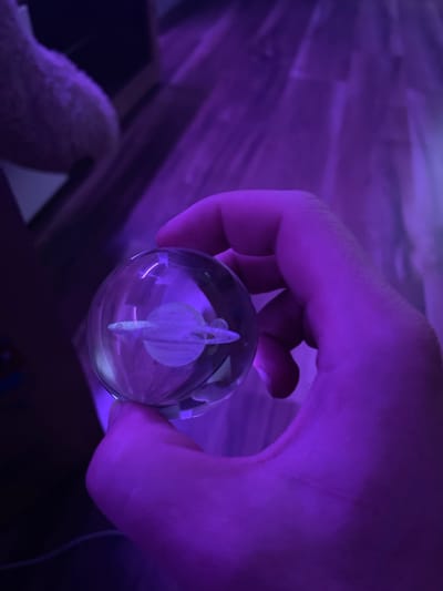 A person's hand is holding a clear glass orb with a miniature Saturn-like planet inside. The orb is illuminated by purple light, creating a vibrant, otherworldly effect. The background is blurred and also bathed in purple light, showing hints of wooden flooring and possibly a plush object. The image is a close-up, focusing on the hand and the orb. The scene is likely indoors, as indicated by the flooring and the artificial lighting. There is no identifiable location in Gliwice, Poland.