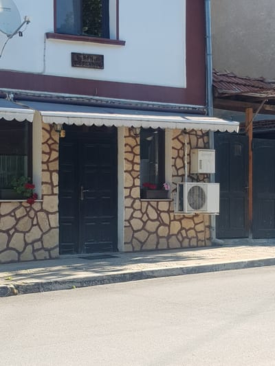 The image depicts the exterior of a building in Lomets, Bulgaria, featuring a facade of stone-like tiles and a dark-colored double door. Above the door, a white awning with scalloped edges is installed. To the left of the door, a window with red flowers in a planter is visible. An air conditioning unit is mounted on the wall to the right of the door. A sign is affixed to the building above the awning, partially obscured by shadow, and appears to have embossed lettering. To the far right, a closed dark gate is present. The building is situated along a paved street, with a concrete sidewalk in front of it. The surrounding environment is sunny, casting shadows on the sidewalk and street.