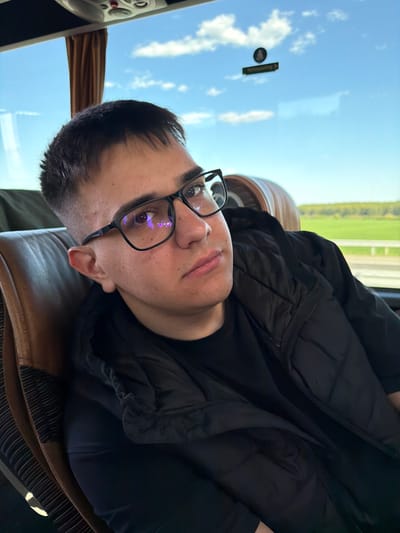 A man wearing glasses and a black jacket is sitting on a brown leather seat, likely inside a vehicle. Through the window, the sky is visible with some clouds. A green field is also seen in the background. The location cannot be determined as Malko Tranovo from the image alone.