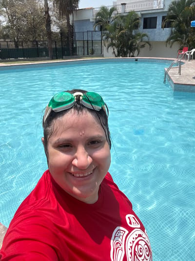 The image depicts a smiling person taking a selfie in a vibrant blue swimming pool under bright daylight in Tres de Mayo, Mexico. The subject, positioned in the foreground, is partially submerged in the water, with their head and upper torso visible. They have wet, dark hair and water droplets on their face, suggesting recent activity in the pool. Green swimming goggles are pushed up onto their forehead. The person is wearing a red long-sleeved shirt adorned with distinctive white tribal-like patterns on the right shoulder and arm. Their wide smile reveals their teeth, conveying a cheerful and happy emotion.
The setting is an outdoor swimming pool, characterized by clear, light blue water reflecting sunlight, with a visible grid pattern from the tiles at the bottom. A light-colored concrete or tiled deck surrounds the pool, where a metallic ladder for access can be seen, along with red and white plastic chairs further back. The background features lush green foliage, including palm trees, partially obscuring a multi-story building with light-colored walls and dark windows. A white fence or railing is also visible, indicating a residential or recreational environment. The bright, clear conditions suggest sunny weather. No discernible text is present, apart from the decorative patterns on the shirt.