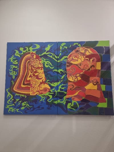 The image displays a diptych artwork mounted on a plain, light-colored wall, likely indoors in Jaunmārupe, Latvia. The artwork consists of two rectangular canvases positioned side-by-side. The left canvas features an abstract profile of a face or head-like shape, oriented to the right. This shape is composed of intricate swirling patterns and bands of color including red, orange, yellow, and brown. It is set against a deep blue background with vibrant, neon green, flame-like or organic swirling patterns emanating from around the central figure. The right canvas mirrors this composition with an abstract profile facing left, also composed of a patchwork of red, orange, yellow, green, and purple hues, and distinct swirling motifs. This figure is similarly set against a deep blue background with matching neon green organic patterns, which appear to connect and intermingle with those from the left panel in the central space between the canvases. The rightmost edge of the right canvas incorporates a checkered pattern in shades of green, blue, and purple. The two central figures appear to be confronting or interacting with each other through their orientation and the connecting green patterns. No people are visible, and no actions or interactions are depicted beyond the static artistic composition. Small, white identification labels are visible beneath the lower edge of each canvas.