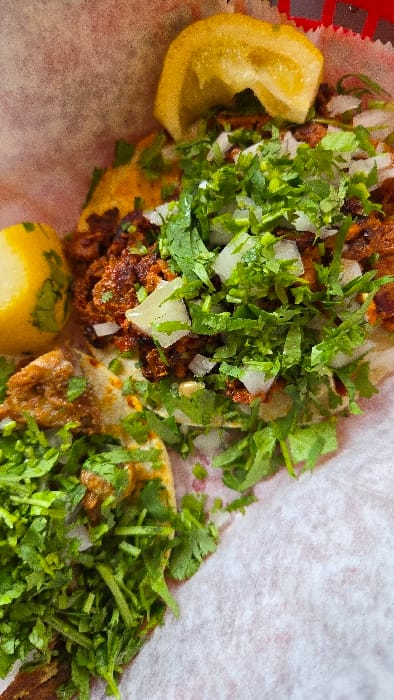 Two tacos filled with seasoned meat, onions, and cilantro sit on a white paper liner. Wedges of lemon garnish the tacos. The meat appears slightly browned and crispy, complementing the freshness of the cilantro and onions. The vibrant colors and textures suggest a flavorful and appetizing meal.