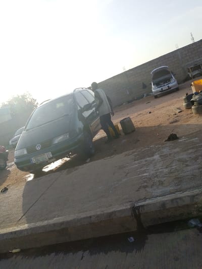 In Gbaza, Nigeria, a man, possibly a mechanic, stands beside a dark green Volkswagen minivan. The van's hood is open, and its windshield appears to have a spiderweb crack. The scene is set in an outdoor area with a dusty, dirt ground and a concrete barrier in the foreground. In the background, a brick wall encloses the area, and a silver car with its hood up is visible. Several old tires are stacked near a yellow container. The sun is low in the sky, casting long shadows and creating a bright, hazy atmosphere.