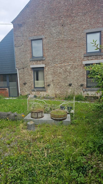 The image depicts an outdoor scene in front of a brick building, likely located in Boussu-lez-Walcourt, Belgium. The building features a dark gray slate roof on the left side and a red brick facade with multiple windows on the right. In the foreground, a grassy area with various plants and weeds is visible. A white decorative metal structure, possibly part of a garden bed or a bench, sits on a concrete base. Two wooden barrel planters filled with grass and small plants are placed on the concrete base. A fallen log and some concrete blocks are also present in the grassy area. The environment appears somewhat overgrown and rustic.