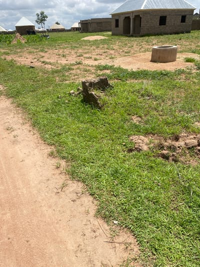 The image shows an outdoor scene in Tavo, Nigeria. The perspective is from a low angle, looking up and slightly to the right. In the foreground, there is a patch of green grass adjacent to a dirt path. A large, rough-hewn rock or clump of earth sits on the edge of the grass.

Further into the scene, a circular concrete structure, possibly a well or water tank, is visible. To the left of the well, there are unfinished brick buildings with corrugated metal roofs, typical of construction in the area. A few other buildings with roofs are visible in the distance, suggesting a developing community. A small, leafy tree stands near some of these structures.

The ground is a mixture of dry, sandy earth and sparse green vegetation. The sky is overcast with white clouds, indicating it might be daytime, though the light suggests it could be morning or late afternoon. There are no people visible in the image. The overall impression is one of rural development in a tropical or subtropical environment.
