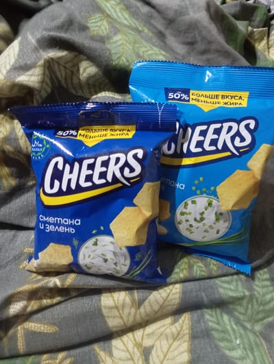Two bags of "CHEERS" brand potato chips are displayed on a patterned gray and green fabric. The bag on the left is closer to the viewer and features the flavor "Smetana i Zelen" (Sour cream and greens). It has a blue background with yellow lettering for the brand name and a white circular image of sour cream dip with green onions and dill. A green and white halal symbol is also visible. The bag on the right is slightly behind and to the right of the first bag. It also has a blue background with yellow lettering and the brand name "CHEERS". A yellow banner across the top of both bags indicates "50% БОЛЬШЕ ВКУСА, МЕНЬШЕ ЖИРА" (50% MORE TASTE, LESS FAT). The fabric on which the bags are placed has a repeating pattern of stylized leaves in shades of green and yellow. The image does not provide any information about a specific location in the city of Nurabad, Uzbekistan.