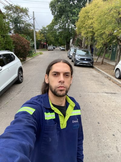 A male individual is depicted in a selfie, standing on a paved residential street in Martínez, Argentina. He has long, dark hair, a beard, and a mustache, and is wearing a dark blue long-sleeved work shirt with a lime green collar and reflective stripes on the shoulders and chest, bearing the "Emova" logo. His expression is neutral, looking directly into the camera. The street is lined with numerous trees exhibiting various shades of green and some reddish foliage. On the left side of the street, a white Volkswagen T-Cross SUV is partially visible, parked facing away from the camera. On the right side, a grey Volkswagen T-Cross SUV is parked, displaying an Argentinian license plate "AE 256 ZV". Further down the street, additional vehicles are parked on both sides, and residential houses are partially visible behind fences and trees. A utility pole with overhead wires is present on the left. The sky is overcast, providing diffuse lighting. No other individuals are visible.