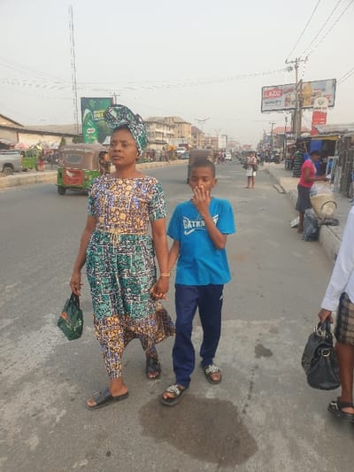 On a busy street, a woman in a patterned dress and headwrap holds hands with a young boy in a blue shirt and navy pants. Behind them are vehicles and buildings, including a billboard advertising Laziz. The woman carries a small bag, and the boy has his hand to his face. A few other people can be seen in the background, including one carrying a purse.