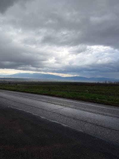 The image shows a view from what appears to be a paved road in Dobroslavtsi, Bulgaria. The road surface is dark and possibly wet. To the left of the road, there is a narrow strip of green vegetation or field. Beyond this, the landscape opens up to a view of a valley or plain with a city visible in the distance. In the far distance, there are mountains or hills. The sky is filled with heavy, overcast clouds, indicating possibly stormy or rainy weather conditions. There are no people visible in the image.