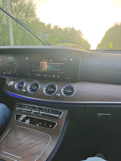 The image shows the interior of a car, likely a Mercedes-Benz based on the dashboard design and air vents. The perspective is from the passenger seat, looking towards the driver's side and the dashboard. The dashboard features a large infotainment screen displaying song information for "Я бы заделался маленьким ко... MiyaGiМаленький Колибри" via Bluetooth. The time displayed is 19:49, and the temperature inside the car is 18°C. The car is in motion, as indicated by the windshield wipers and the sunlit exterior visible through the windshield. The weather appears to be partly sunny, with water droplets on the windshield suggesting recent rain or mist. The sun is low in the sky, casting a warm glow and creating lens flare, indicating it is either early morning or late afternoon. The interior of the car has wood-grain trim and ambient blue lighting, suggesting a luxurious setting. The center console has climate control buttons and a cubby with a "RADIO MEDIA" button. A person's leg, clad in blue jeans, is visible on the left side of the frame, and another person's foot in a dark shoe is visible at the bottom right, confirming the presence of at least two people in the car. The scene suggests a drive through a rural or suburban area, possibly on the outskirts of Репниково, Russia, given the context. The overall mood is relaxed and everyday, with the focus on the car's interior and the music playing.