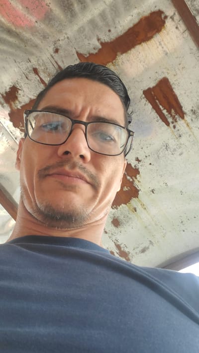 A close-up, low-angle portrait shows a man looking down directly into the camera. He has short, dark hair, black-framed glasses, and a short goatee or stubble. He is wearing a dark blue t-shirt. His expression is serious or intent, with a slightly furrowed brow. The image is rotated, presenting the man's face at an angle. The background behind him is a textured, light-colored surface, possibly metal or concrete, extensively marked with prominent brown, rust-like stains and specks. This weathered surface appears to be a ceiling or an overhead structure. The lighting suggests it is daytime. There is no visible text in the image. The location context is El Papelón, Venezuela.
