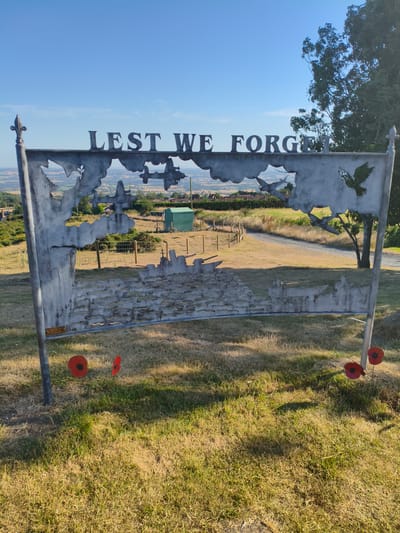 The image shows a large metal memorial sculpture on a grassy hill. The sculpture is shaped like a banner and features cut-out silhouettes of World War II aircraft, bombing raids, and doves. The words "LEST WE FORGET" are prominently displayed in raised metal lettering at the top of the sculpture. In the foreground, several artificial red poppies are placed on the grass.
The background reveals a rural landscape with rolling hills, fields, and scattered houses, suggesting an elevated position. The sky is clear blue with a few wispy clouds. The overall scene evokes a sense of remembrance and patriotism. The memorial is located in Cleehill, United Kingdom.