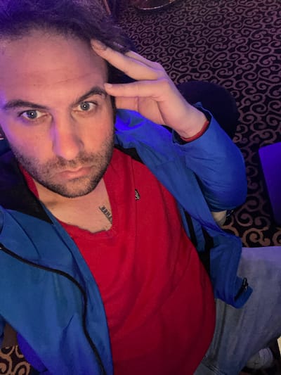 A seated adult male is captured in an indoor setting within the city of Armagh, United Kingdom. He looks directly at the camera with an intense gaze, his left hand resting on his forehead, partially obscuring his dark, textured hair. He has blue eyes and a dark beard/stubble. He is dressed in a red collared shirt, visible beneath a blue zippered jacket featuring black panels along the sides. Light gray trousers are visible from the waist down. A red string or bracelet adorns his right wrist. A black text tattoo, reading "THER" (inverted from the viewer's perspective), is partially visible on his upper right chest. The environment is dimly lit with a blue/purple ambient hue, revealing a dark carpet patterned with intricate, swirling dark brown and black motifs. A dark, indistinct object is present to the lower right of the frame.