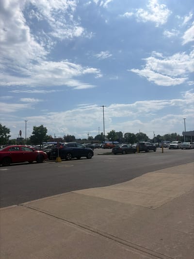 The image depicts a parking lot at a large retail establishment, likely a big-box store, in Longueuil, Quebec, Canada. The parking lot is asphalt and shows some wear and tear with visible lines demarcating parking spaces. Several vehicles are parked in the lot, including a red sedan in the foreground, a dark blue SUV, and other cars further back. Some trees and streetlights are scattered throughout the lot. In the background, there are buildings, likely commercial, and power lines. The sky is partly cloudy with a bright blue sky visible. The overall scene suggests a typical sunny day in a suburban commercial area.