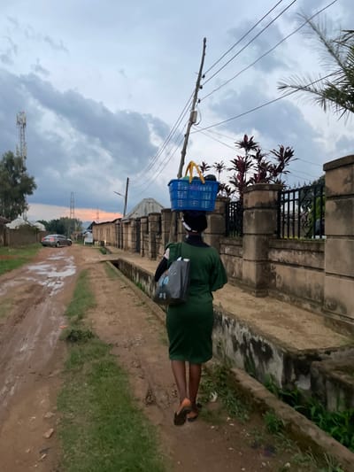A woman walks along a dirt road in Barikin Jirga Summa, Nigeria, carrying a blue basket on her head filled with goods. She is wearing a green dress and carrying a grey bag in her left hand. The road is muddy and uneven, with puddles of water visible. On the right side of the road, there is a concrete wall with a black metal fence. Behind the wall, there are plants and buildings. In the background, there is a cell tower and trees. The sky is cloudy, and the lighting suggests it is either early morning or late evening.