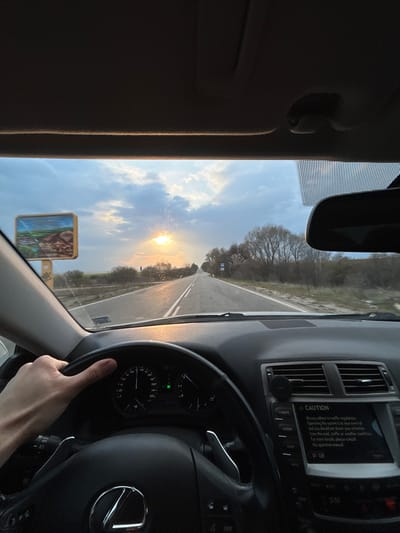 The image captures a first-person view from the driver's seat of a Lexus vehicle on a two-lane asphalt road in Grigorevo, Bulgaria, during the late afternoon or early evening. A left hand is visible gripping the black leather steering wheel, centered on the lower-left quadrant of the frame. The dashboard is visible, featuring an instrument cluster with a speedometer needle indicating approximately 60-70 units (likely km/h) and a tachometer. To the right of the steering wheel, the infotainment screen displays a "CAUTION" message regarding traffic regulations and not diverting attention. Air vents are visible on the dashboard, and a phone mount is affixed to the vent on the right.

The windshield is partially obscured by sun glare and numerous small specks, possibly dust or water droplets. Beyond the windshield, a straight road extends into the distance under a partly cloudy sky with a low-hanging sun on the horizon, casting a golden light and significant glare directly towards the vehicle. The sun's light creates sun rays extending into the clouds. On the left side of the road, a large billboard with a landscape image (depicting green hills and water) is visible. The roadside features bare trees and sparse brown vegetation, indicative of a dormant season or early spring. Further down the road on the right, additional road signs are discernible, but their text is indistinct. A rearview mirror is visible in the upper right portion of the windshield. The interior roof lining is dark.