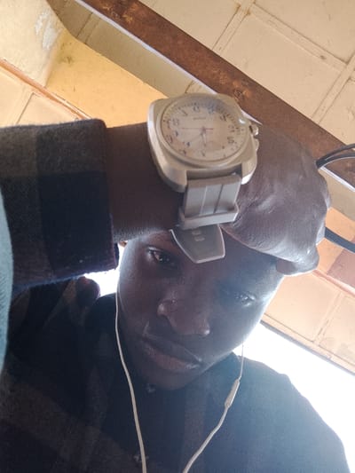 A low-angle selfie depicts a dark-skinned individual in an indoor setting, identified as the city of Toro, Nigeria. The person looks directly into the camera, wearing a dark plaid long-sleeved shirt. A large, light-colored wristwatch with a matching, ribbed strap and Arabic numerals is prominent on their left wrist. White wired earphones are visible, with one earbud positioned in the left ear. The background consists of an overhead ceiling structure featuring light-toned panels with visible peeling paint and dark, discolored horizontal beams.
