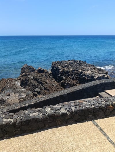 The image presents a serene coastal landscape in Puerto Del Carmen, Spain, captured on a bright, clear day. The upper portion of the frame features a vast, light blue sky with no clouds, meeting a distinct horizon line where it merges with the ocean. The sea itself displays various shades of blue, from a darker hue near the horizon to a more vibrant turquoise closer to the shore, with subtle ripples indicating calm waters.
In the mid-ground and foreground, a rugged coastline is visible, characterized by dark, porous volcanic rock formations that extend into the sea. These rocks are irregular in shape and texture, suggesting a natural geological feature. Along the edge of a paved area, two parallel structures resembling low walls or raised borders are constructed from similar dark, irregular volcanic stones, blending with the natural environment.
The immediate foreground reveals a light-colored, pebbled pavement, likely a pathway or promenade, offering a textured contrast to the darker rocks. A decorative strip of darker, smaller stones is integrated into this pavement. No human activity or specific events are depicted, nor is any visible text present. The scene conveys a sense of tranquility and natural beauty, highlighting the unique volcanic geology against the backdrop of the clear sky and calm sea. The bright sunlight suggests it is daytime, with shadows indicating a high sun.