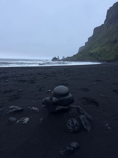 The image depicts a black sand beach in Vik, Iceland. A rugged, moss-covered cliff rises dramatically on the left. A grey sky covers the top left corner. The Atlantic Ocean is visible as a strip of white waves along the left, meeting the black sand. Several smooth, dark grey stones are clustered together in the foreground on the beach, some with light-colored markings. Footprints and smaller rocks are scattered across the dark sand.
