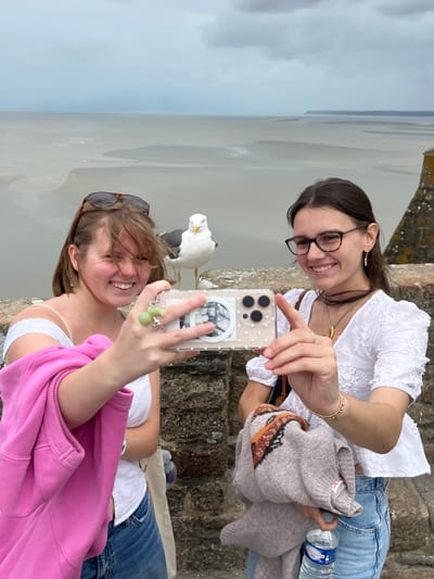 In the city of Le Mont-Saint-Michel, France, two young women are taking a selfie with a seagull perched on a stone wall between them. The woman on the left, with blonde hair and sunglasses perched on her head, smiles brightly as she holds a smartphone. She wears a white tank top and a pink hoodie slung over her arm. The woman on the right, with dark hair and glasses, also smiles and points towards the phone. She is dressed in a white, embroidered top and blue jeans. Behind them, the vast, grey expanse of the bay of Mont-Saint-Michel stretches out under an overcast sky. The stone wall in the foreground and the distant landscape suggest a historic and scenic location.