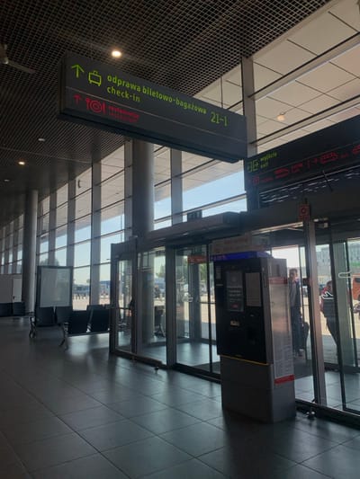 The image shows the interior of a modern airport terminal. A large sign with white text on a green background is located in the center of the frame at the top. The sign reads "odprawa biletowo-bagazowa 21-1 check-in" and "restauracja restaurant". There is also a smaller sign above it with a white arrow pointing up and a suitcase icon. The sign is positioned above a set of glass doors leading to the outside. There is another sign on the right with a red arrow pointing to the right and the words "wyjście exit" in white. This sign is next to a large, modern, black and gray parking machine, facing to the right. Two people are standing to the right of the parking machine. The floor is tiled in gray and black. There are rows of seats with a black frame and light gray cushions. The large windows provide views of the outside. The environment is modern and spacious, with a sense of order and efficiency. The image was taken at Pyrzowice Airport in Pyrzowice, Poland at latitude: 50.471, longitude: 19.073.