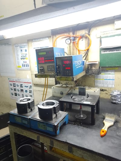 This image depicts a workshop or manufacturing setting in Ciantra, Indonesia. The scene is dominated by industrial equipment and materials, suggesting a focus on production and quality control.

In the foreground, two large, metallic clamps are positioned on blue platforms. These appear to be part of a larger machine or testing apparatus. A black metal table with several small components, including what looks like a stamping tool and pins, sits to the right of the clamps. A paintbrush with an orange handle and white bristles rests on the workbench to the right, hinting at potential cleaning or finishing activities. In the background, there are two digital measurement devices with red and green displays, labeled "TOSOK". One displays "822.5" and "123.5", while the other shows "70.31". Above the digital displays, a small pressure gauge with an orange hose is visible, connected to a pipe.

Several posters or charts are affixed to the wall. One, titled "INSTRUKSI KERJA" (Work Instructions), is positioned above the measurement devices. Another, featuring diagrams of various machine parts and labeled "TOSOK" with Indonesian text, is on the left. A green board with text that includes "TEMPAT LAPORAN HARIAN" (Daily Report Location) is on the right. There's also a document labeled "MONITORING CUSTOMER CLAIM" on the left side of the wall.

The lighting suggests an indoor environment, possibly with artificial lights as indicated by the bright ceiling fixtures. The overall impression is one of a functional, albeit somewhat cluttered, industrial workspace. The visible text, primarily in Indonesian, confirms the location and hints at the operational context, including work instructions, daily reporting, and customer claim monitoring. There are no people visible in the image.