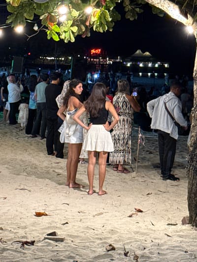 The image depicts a nighttime scene on Cenang Beach, Malaysia, at what appears to be a lively gathering or event. In the immediate foreground, string lights with incandescent bulbs illuminate a sandy beach strewn with scattered dry leaves and debris. A tree trunk is partially visible on the far right.

In the midground, a crowd of individuals stands on the sand, generally facing away from the camera toward a lit background area. Two women are prominent in the center. The woman on the left has dark, curly hair, wears a light-patterned top and light shorts/skirt, and carries a light shoulder bag. Her hands are on her hips, and she is seen in profile facing left. The woman on the right has dark, straight hair, wears a black top and a white pleated skirt, and also has her hands on her hips, facing away from the camera. Both women appear to be barefoot or wearing minimal footwear.

Behind them, a dense line of people extends across the frame. Visible figures include an individual with light pink hair holding a phone to capture an image on the far left, several people in dark attire, and a woman in a long, patterned dress. On the far right, a man in a light grey hoodie and dark track pants with white stripes is visible, carrying a dark shoulder bag.

In the background, a brightly lit area suggests establishments or event structures. A prominent red neon sign reads "The CL?T" (with the '?' possibly being an '@' or other character). Additional background lights illuminate distant tents or buildings against the dark night sky.