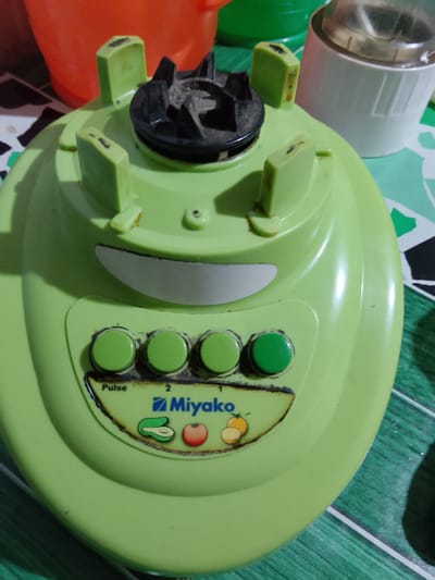 The image shows a close-up, high-angle view of a light green blender base with four green circular buttons on the front. The buttons are labeled "Pulse", "2", and "1". Below the buttons, a logo with the brand name "Miyako" is visible, along with icons of an avocado, a tomato, and two orange slices. The blender base has a curved indentation above the buttons, resembling a smiling mouth. In the background, out of focus, are parts of orange and green plastic containers, and a white container with a clear lid. The blender base rests on a surface with a green and white checkered pattern. The scene is indoors, with artificial lighting casting shadows. The image is taken in Merbopuntong, Indonesia.