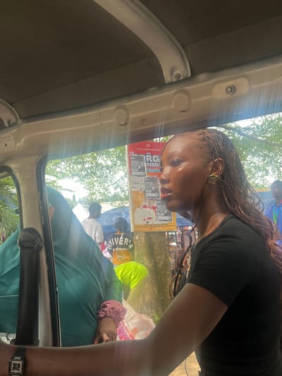 The image, taken from inside a vehicle in Ijanikin, Nigeria, shows a woman with braided hair wearing a black shirt and gold earrings. She is leaning forward, seemingly looking out the vehicle. Outside, there are people, including someone wearing a neon yellow top and a shirt with "RIVER" on it. Various posters are visible in the background. The inside of the vehicle is partly visible, including the doorframe and a seat, where a person in a green shirt and someone with a pink scrunchie on their wrist is sitting. The time shown on the watch worn by the person in the green shirt is 2:15. Trees and buildings are in the distance.