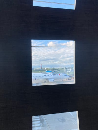 The image depicts an Air France airplane seen through a window. The location is identified as Le Mesnil-Amelot, France, implied but not explicitly stated in the image. The sky is visible in the background, indicating an outdoor setting, likely an airport terminal. No people are directly visible.