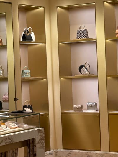 The image depicts an interior view of a luxury handbag store, likely located in Nong Prue Subdistrict, Thailand, given the provided context. The store features brightly lit, gold-accented shelving units displaying a variety of designer handbags. On the top shelf of the left unit, two handbags are visible: one black and white, and the other appears to be a white tote. Below this, on the second shelf, is a small, light green, sequined handbag. The third shelf down shows a light pink quilted handbag. On the bottom shelf of this unit, a black handbag with gold handles is partially visible.
The middle shelving unit displays handbags on its upper shelves. The top shelf has a small, patterned handbag with silver handles. The shelf below it showcases a heart-shaped black and silver clutch with a gold chain handle. The third shelf from the top features two small, rectangular handbags; one is beige and the other is silver.
The right shelving unit is largely out of focus, but a portion of a handbag is visible on the top shelf, appearing to be a small, light brown clutch. In the foreground, a marble-topped display counter with glass is visible, showcasing jewelry on velvet cushions. The overall environment suggests a high-end retail setting with immaculate presentation. No people are visible in the image.