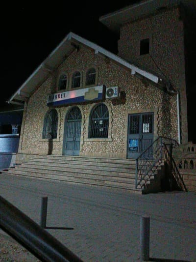 The image shows the facade of a market building at night, located in Copăceni, Moldova. The building is constructed with rough-hewn stone and features a prominent pitched roof with white trim. Above the entrance, a sign with "MARKET" written in white capital letters on a blue background is visible. The main entrance is a large, dark-colored double door with metal grates, accessed by a set of concrete stairs with an ornate black metal railing. To the left of the main entrance, there are arched windows with similar metal grates. An air conditioning unit is mounted on the stone wall above and to the left of the entrance. To the right of the building, a tall, square tower-like structure is attached, also made of stone, with a dark rectangular opening. The foreground shows a paved area with two short, cylindrical posts and a section of a dark metal railing. The overall scene is dimly lit, suggesting nighttime, with the building illuminated by artificial light sources. The environment appears to be an urban setting, given the building's design and the paved area.