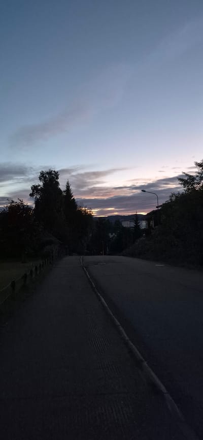 The image captures a twilight scene on a paved road in Lillehammer, Norway. The road curves to the right, bordered by a dark, grassy verge and trees. A subtle white line marks the edge of the road. In the distance, silhouetted trees and houses suggest a residential area. A streetlamp stands near the center of the frame, illuminating the twilight sky.

The sky is a dominant feature, with wispy clouds stretching horizontally. The colors in the sky transition from a pale blue on the left to warmer hues of pink and orange near the horizon, indicating the setting sun or pre-dawn light. Mountains are faintly visible in the far distance, their shapes softened by the atmospheric conditions. The overall mood is calm and serene, typical of the transition between day and night. The time of day is clearly evening or early morning. There are no people or animals visible in the image. No text is present.