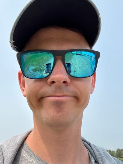 This is a close-up, vertically oriented selfie of a person's face, taken outdoors. The subject is wearing a dark baseball cap with a slightly frayed brim and black sunglasses with bright blue, mirrored lenses. The sunglasses reflect a grassy area and what appears to be a light-colored building or structure. The person's face is angled towards the camera, with their mouth slightly open in a relaxed expression. Light stubble is visible on their chin and jawline. They are wearing a gray t-shirt, the fabric of which is visible on the right side of the image. The background is a plain, light blue sky, suggesting it is daytime. There is no visible text in the image. The overall impression is casual and informal, possibly taken during a sunny day in an outdoor setting like a park or field.