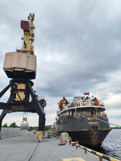 In Shal'skii, Russia, a large industrial crane with a grabber bucket is positioned on a gravel-covered dock. The crane is yellow and black, with a substantial structure supporting a large, box-like operator's cabin. The grabber bucket is suspended from the crane, appearing to be in the process of collecting or depositing gravel. In the background, a dark-hulled ship with a Russian flag flying from its stern is docked. The ship has orange life preservers visible on its deck and the name "ШАЛА" and "МЕДВЕЖЬЕГОРСК" inscribed on its stern. The water appears calm. The sky is overcast with dramatic, dark clouds. The overall scene depicts a port or industrial loading area.