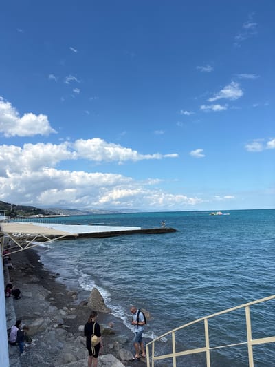 The image depicts a sunny day at a beach in Alushta, Ukraine. The foreground shows a stony beach with people walking and sitting on the rocks. A white staircase with yellow railings leads down to the water. The sea is a vibrant blue, with small waves lapping at the shore. In the midground, a concrete pier extends into the sea, and a few people are visible on it. Further out, a floating water park with colorful inflatables can be seen, along with a yellow boat. The background features a coastline with buildings and hills under a bright blue sky dotted with fluffy white clouds.