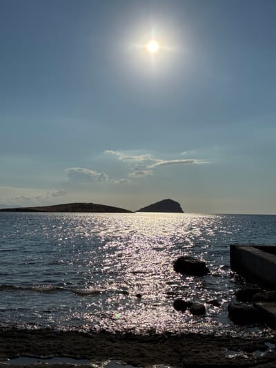 A sun glare over the Aegean Sea is captured in this vertical, outdoor, full shot of a seascape. The sun is positioned in the upper center of the frame, its rays illuminating the water with a brilliant sparkle. In the middle ground, two islands are visible in silhouette against the hazy horizon. The island on the left is low-lying and extends horizontally, while the island on the right is a more prominent, conical shape with a small structure at its peak. The foreground features a rocky shoreline with a concrete structure extending into the water on the right. The water is a deep blue, reflecting the sun's light in a shimmering pattern. The sky is a clear, pale blue with a few wispy clouds. The scene appears to be located in the vicinity of Limenas Markopoulou, Greece, given the characteristic island formations and the Aegean Sea. There are no people or animals visible in the image.
