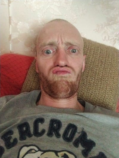 A close-up, high-angle selfie shows a bald, light-skinned man with a red beard making a silly face. He has a light-colored sweatshirt with dark lettering, some of which spells out "ABERCROMBIE" with a graphic of a dog underneath. The man is leaning against a textured brown couch, with a red pillow visible to his left. The background consists of a wall with a floral wallpaper pattern. The man's facial expression is one of exaggerated surprise or displeasure, with his eyebrows furrowed and his mouth puckered. The lighting is from above and to the right, casting some shadows on the man's face.