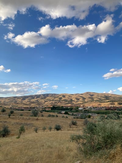 The image depicts a wide-angle view of a landscape near Avanos İlçe Merkezi, Turkey, on a clear day with scattered white clouds. In the foreground, dry, golden-brown grass and sparse, small, scrubby trees cover rolling hills. A dirt road is visible winding through the terrain. In the mid-ground, a small settlement with terracotta-roofed buildings and some industrial structures is nestled amongst a strip of greener vegetation. Beyond the settlement, a range of undulating, dry, golden-brown hills forms the background. The sky is a vibrant blue. The overall impression is one of a rural, arid environment.